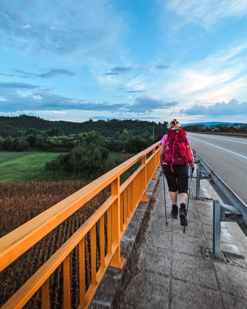 Female pilgrim wearing a red backpack and with hiking poles, walking on the side of the street with a yellow railing