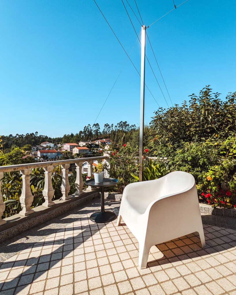 White chair and small metal table on a balcony surrounded by plants and hillside views at Albergue Peregrinos St Antonio de Agueda