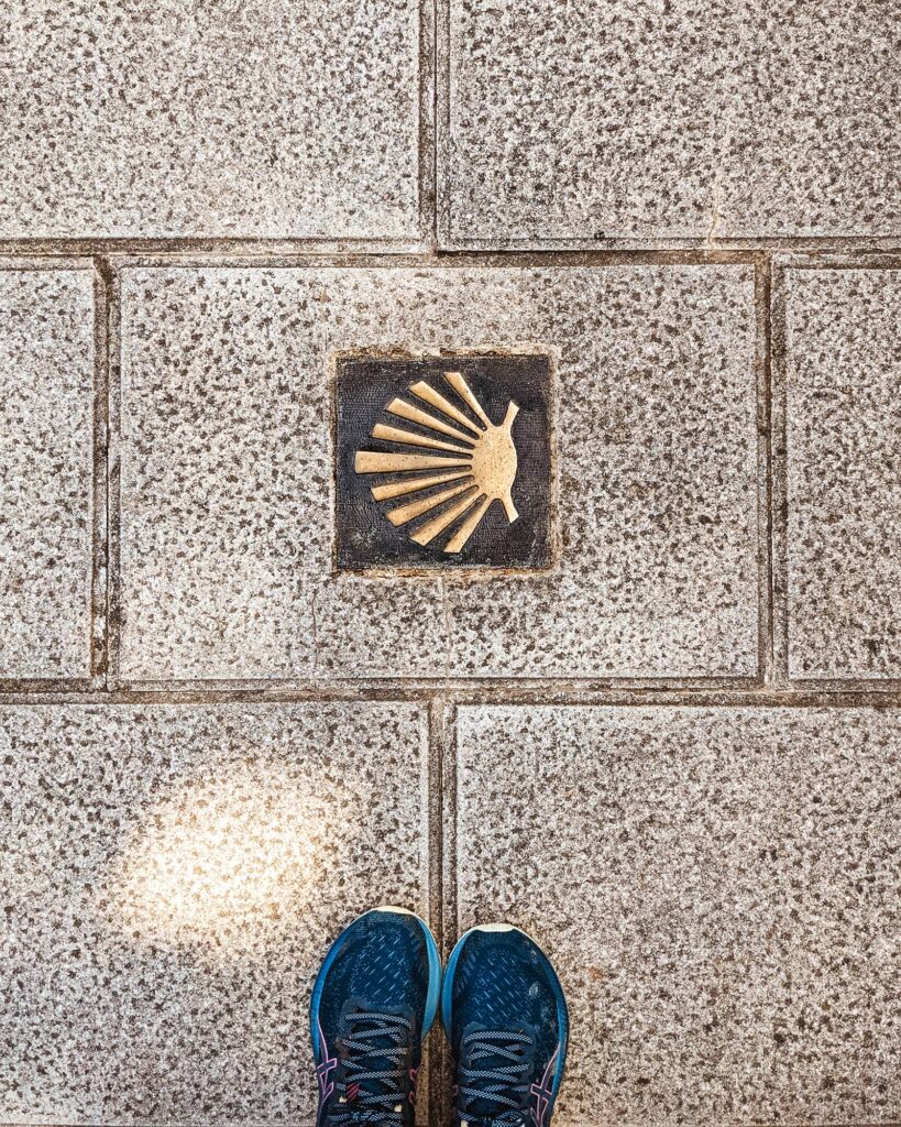 Top down view of blue ASICS running shoes and the Camino de Santiago gold shell marker on a stone street