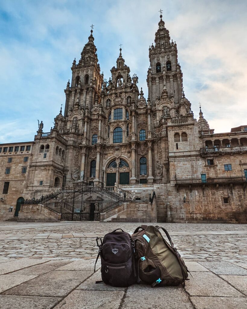 A black Osprey backpack and a gray Osprey backpack sitting on the ground in front of the Cathedral of Santiago de Compostela