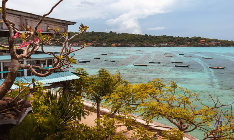 View of trees, a wooden building, and a sand path in front of turquoise blue clear water with boats and an island in the background