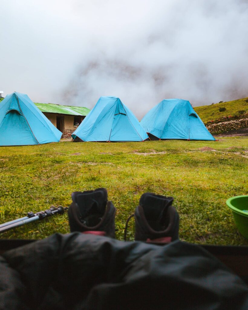 View from inside a tent on the Salkantay trail of hiking boots, hiking poles, a green bowl of water, grass, blue tents, and fog