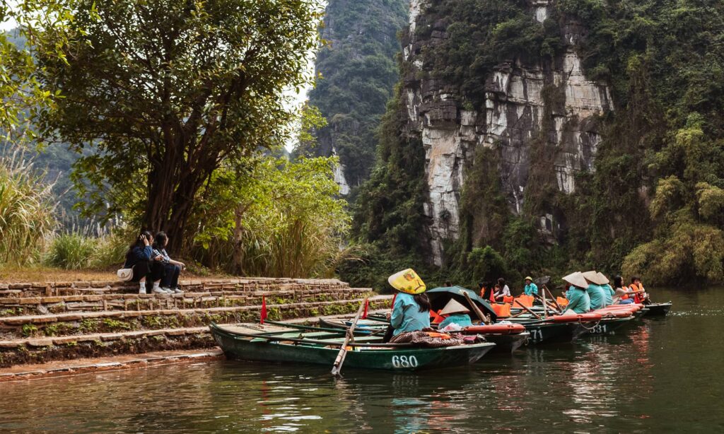 Rowboats with local guides in traditional conical hats line the riverbank in Trang An, a must-visit destination on any Ninh Binh itinerary, with dramatic limestone cliffs and lush forest creating a scenic backdrop.
